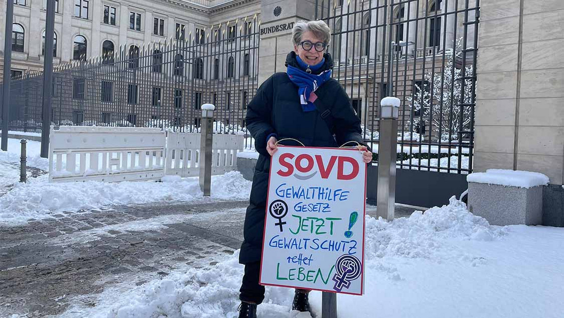 Frau steht vor dem Gebäude des Bundesrats. Sie trägt ein Plakat mit der Aufschrift "Gewalthilfegesetz jetzt!"