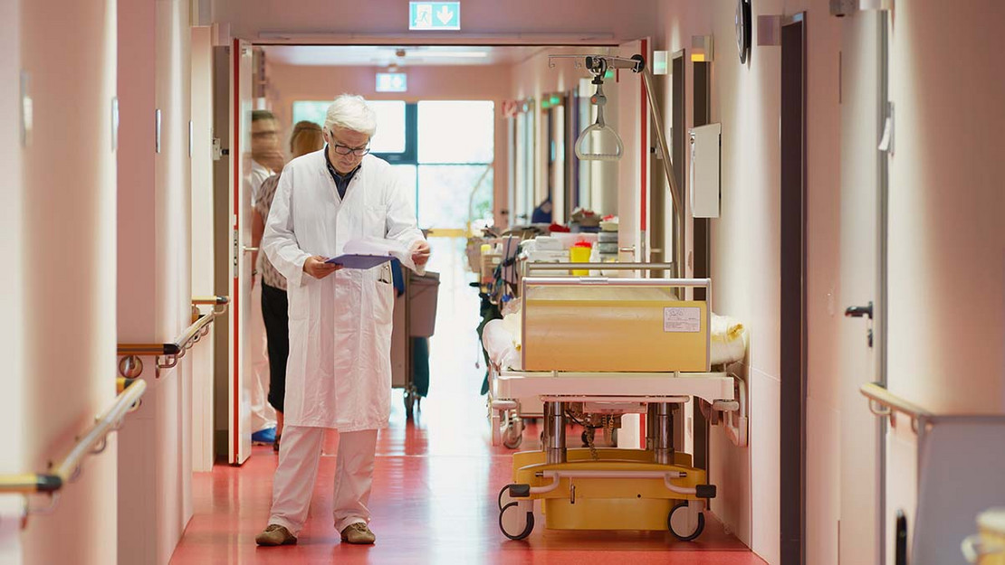Älterer Arzt steht im Gang eines Krankenhauses und liest in Unterlagen.